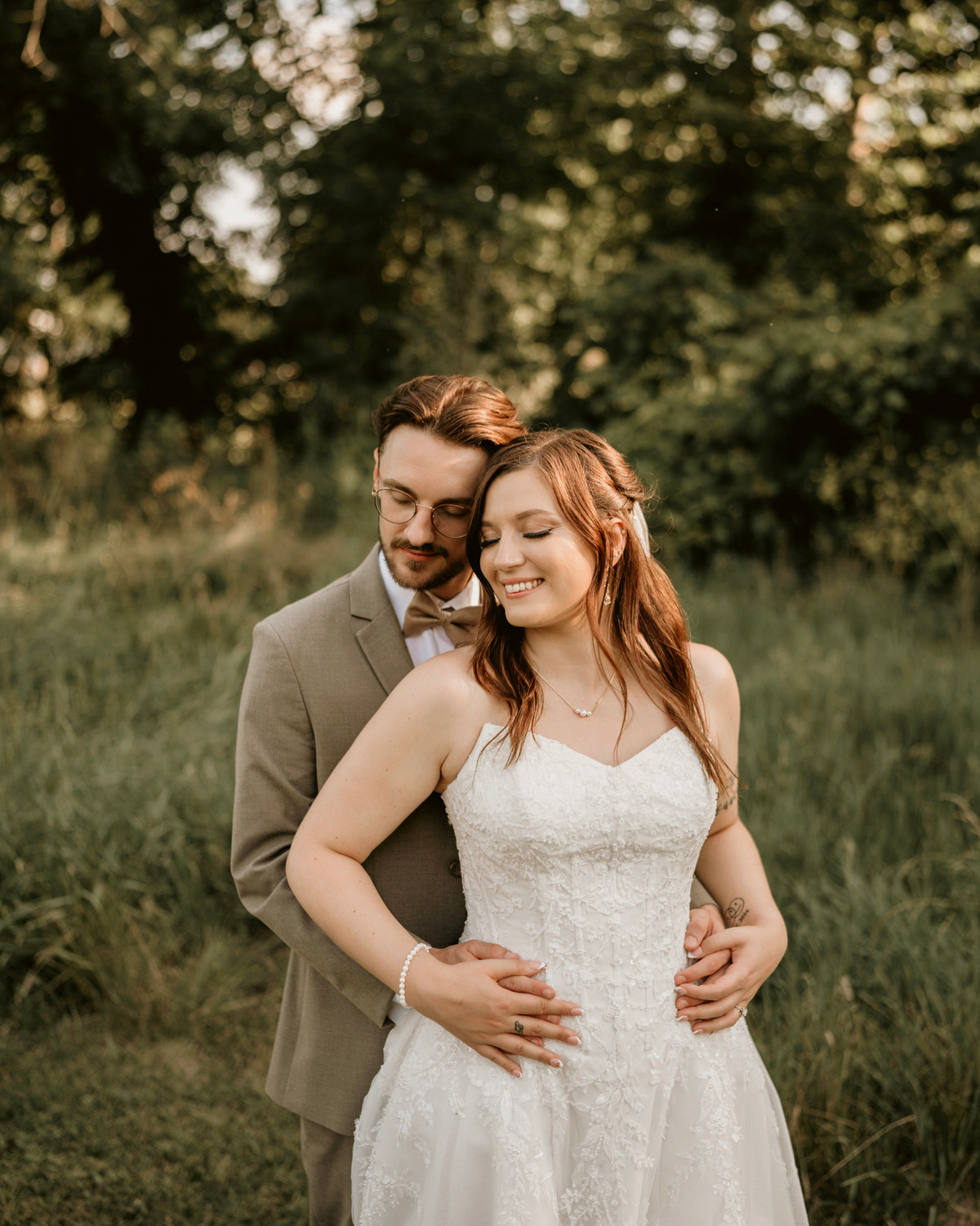 Groom in a taupe suit embracing his smiling bride from behind in a sunlit meadow