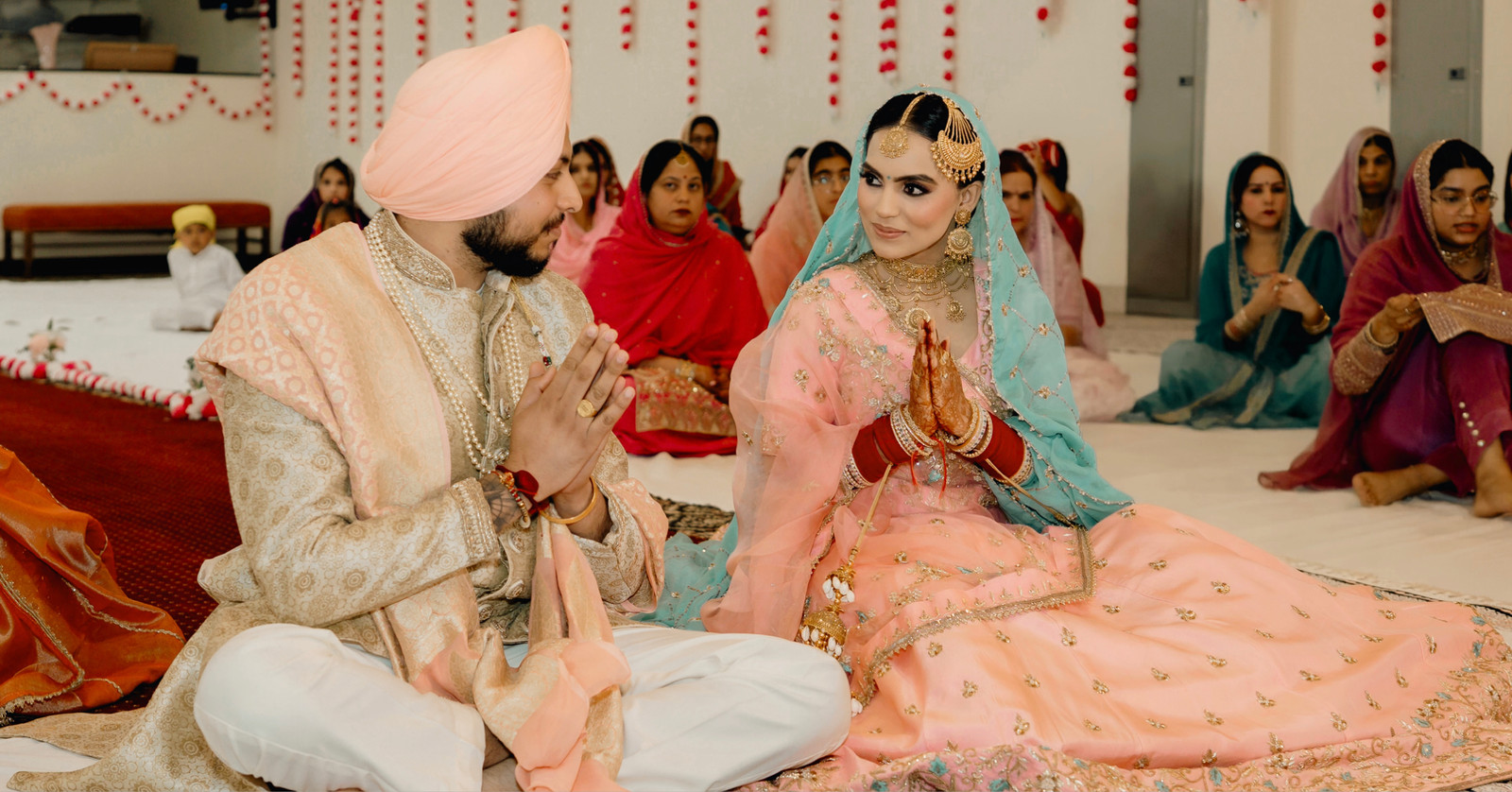 Sikh bride in peach and turquoise and groom in a peach turban sitting with folded hands during their Anand Karaj, family seated behind them