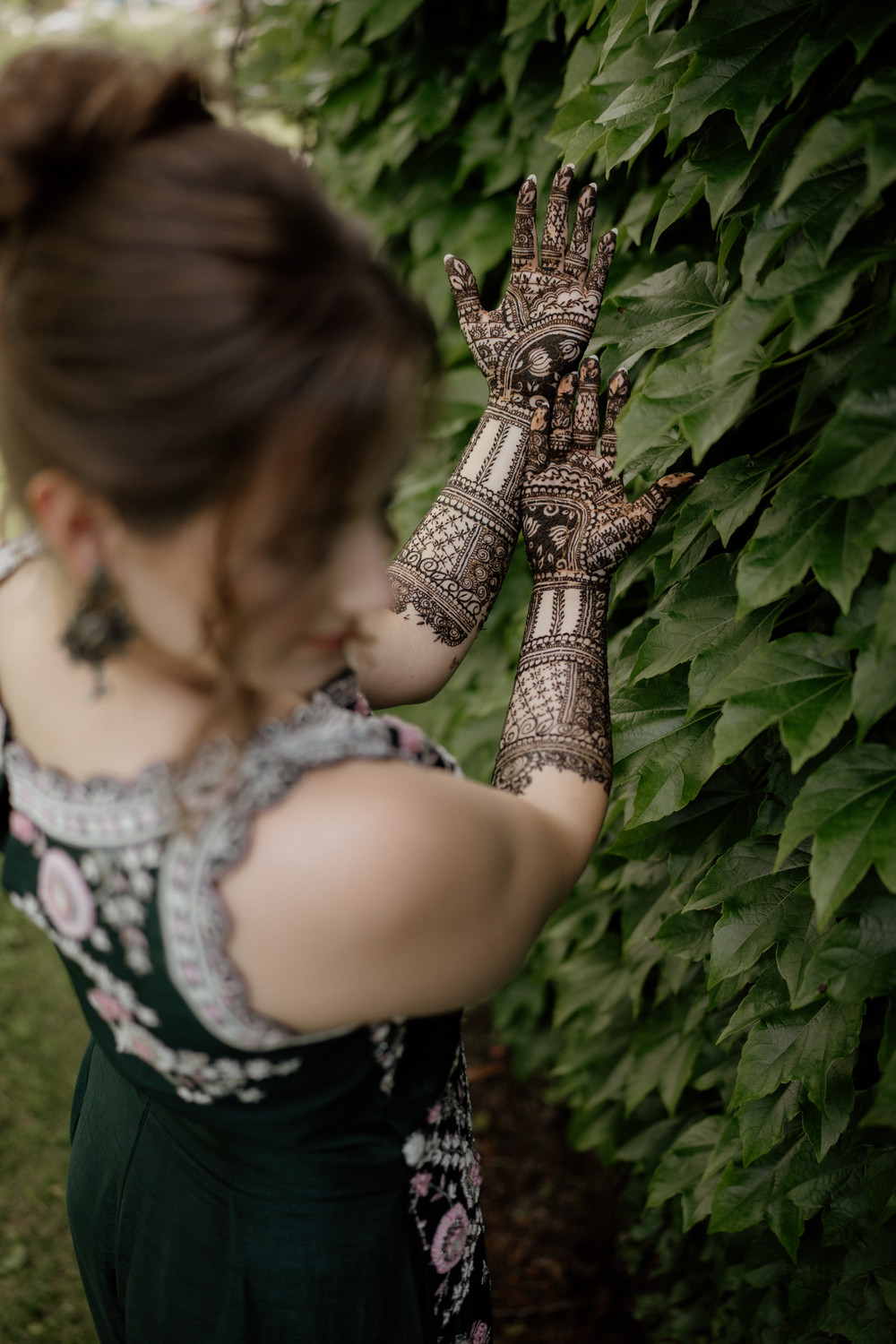 Bride raising both hands covered in intricate dark mehndi against a wall of green leaves