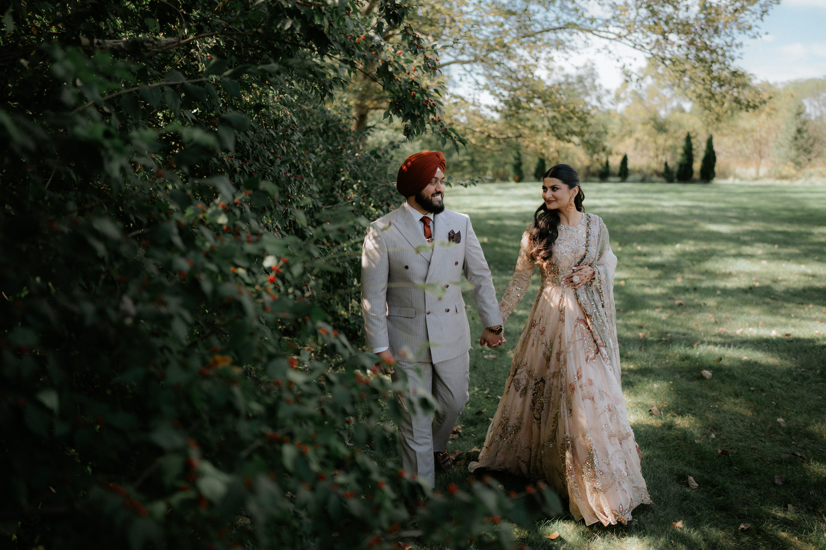 Bride in a blush embroidered lehenga and groom in a grey suit and rust turban walking hand in hand through a sunlit garden