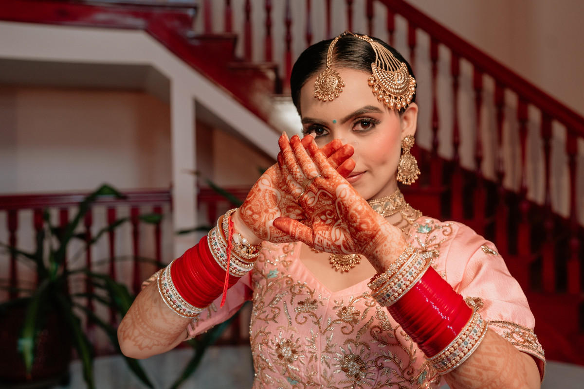 Bride in a peach lehenga and gold matha patti peeking over her hennaed hands, red chooda on her wrists