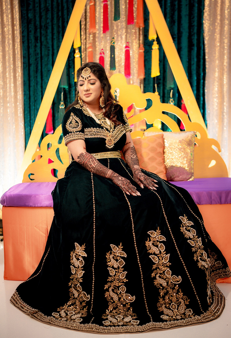 Bride in a dark green velvet outfit with gold embroidery and full mehndi, seated on a colorful mehndi stage