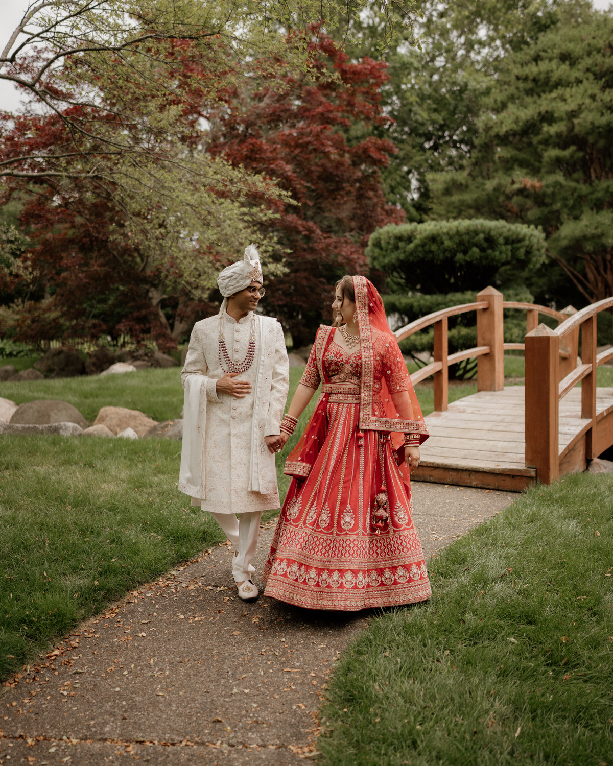 Bride in a red embroidered lehenga and groom in an ivory sherwani and safa walking hand in hand past a wooden garden bridge