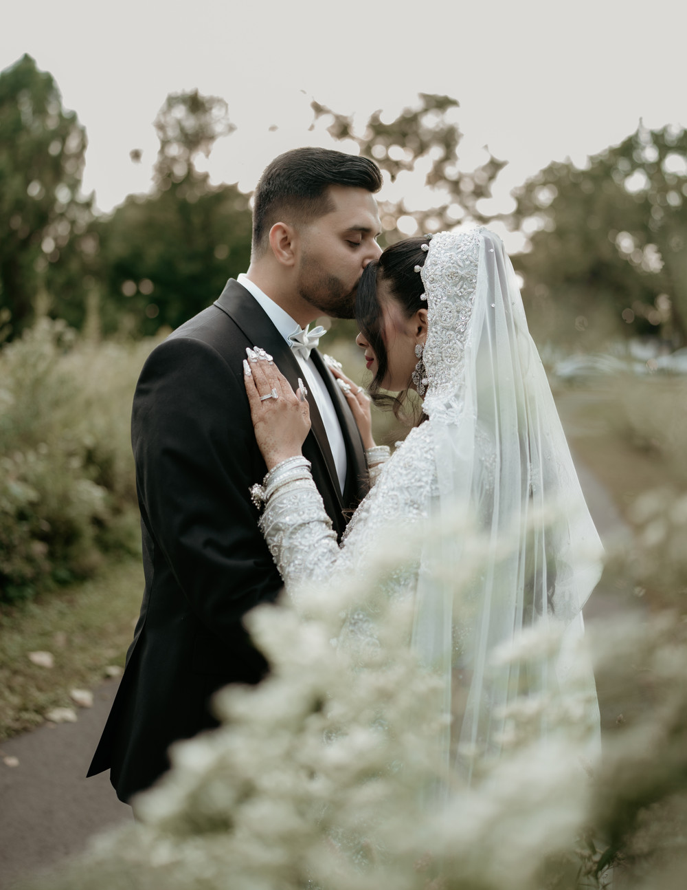 Groom in a black tuxedo kissing the forehead of his bride in a white embellished outfit and veil