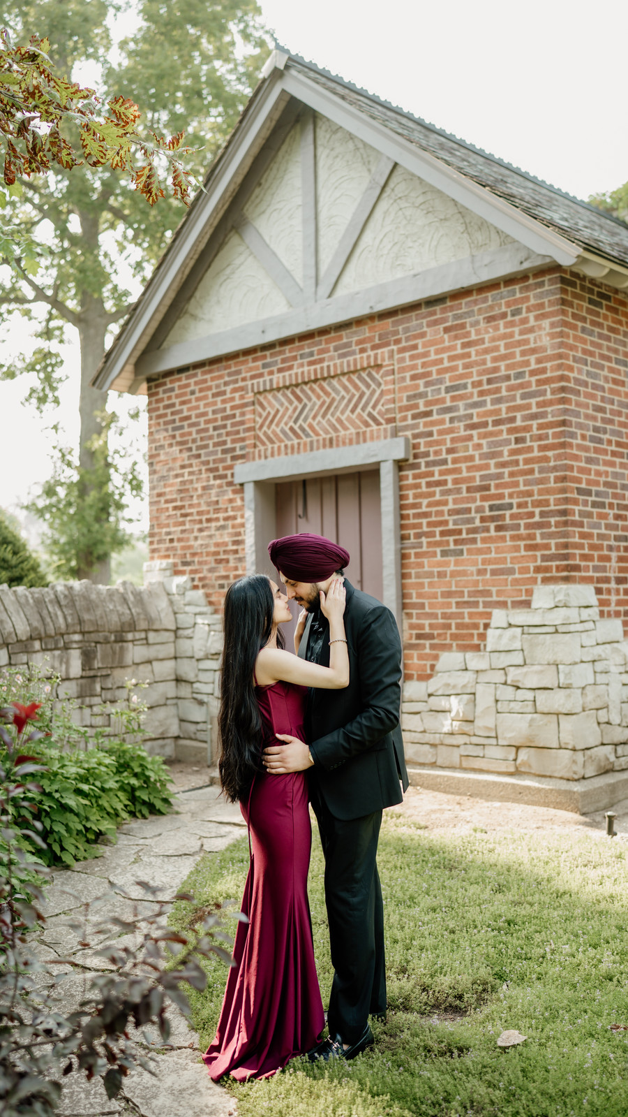 Couple in a burgundy gown and black suit with a maroon turban, about to kiss beside a red brick cottage
