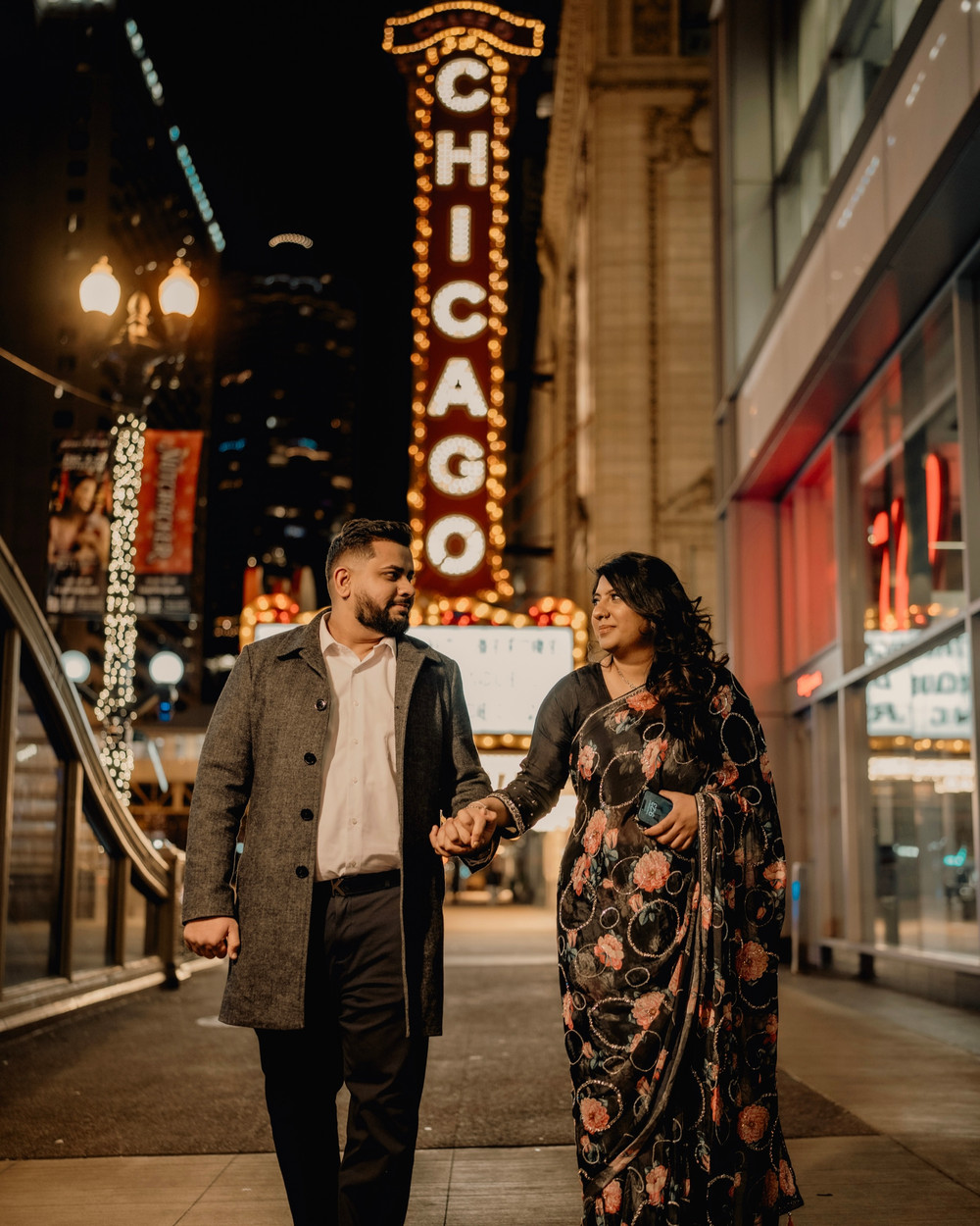 Couple walking hand in hand at night beneath the lit Chicago Theatre sign, she in a floral saree and he in a grey overcoat